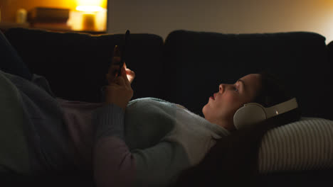 Mujer-Con-Auriculares-Inalámbricos-Recostada-En-Un-Sofá-En-Casa-Por-La-Noche-Transmitiendo-O-Mirando-Contenido-En-Línea-En-Un-Teléfono-Móvil-3